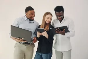 Three people smiling and looking at their devices while using tablets and a laptop