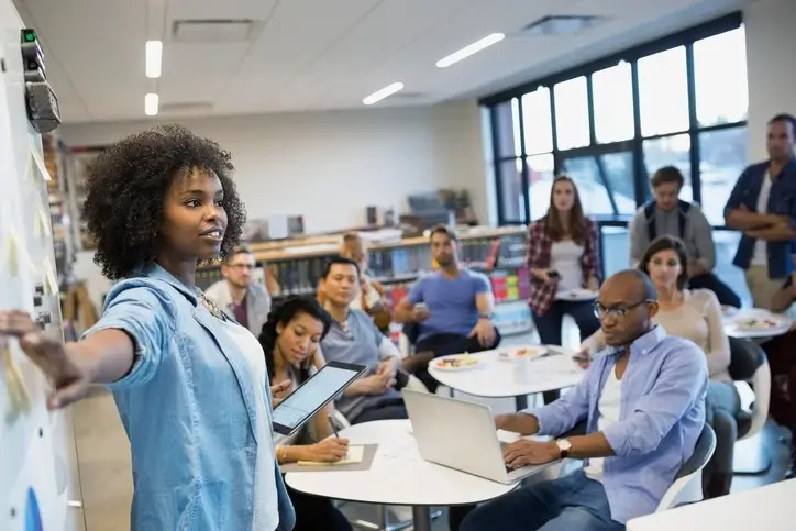 A woman standing at a whiteboard pointing to an illustration while speaking to a group of people